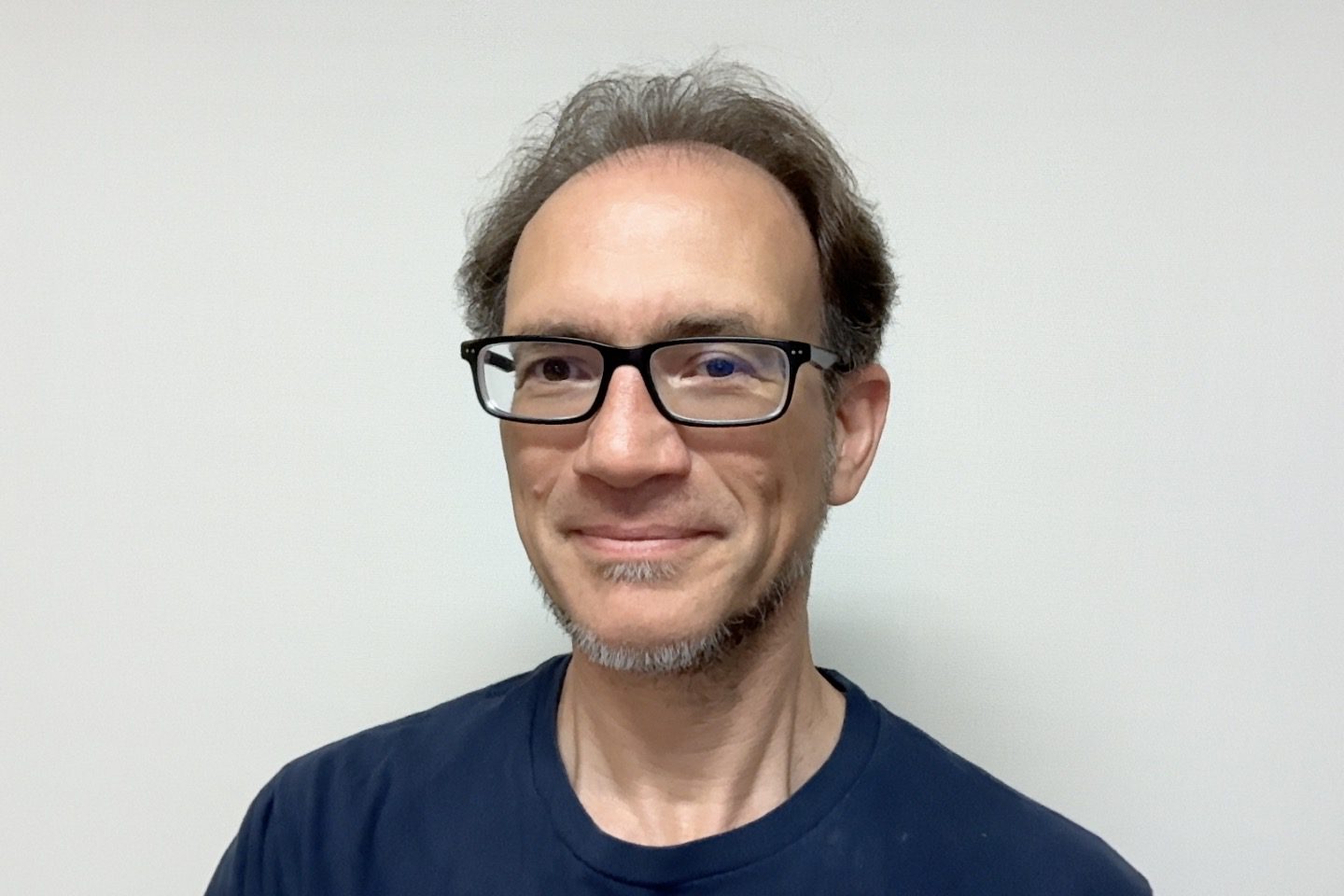 A middle-aged man with glasses, short brown hair, and a trimmed beard wears a dark T-shirt and smiles at the camera against a plain light background.