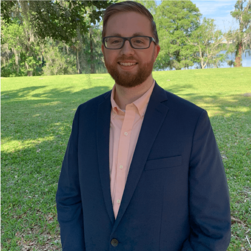 A man with glasses and a beard stands outside wearing a navy suit and peach shirt. Trees and grass are in the background.