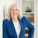 A woman with gray hair wearing a blue blazer and white shirt stands indoors, smiling, with bookshelves in the background.