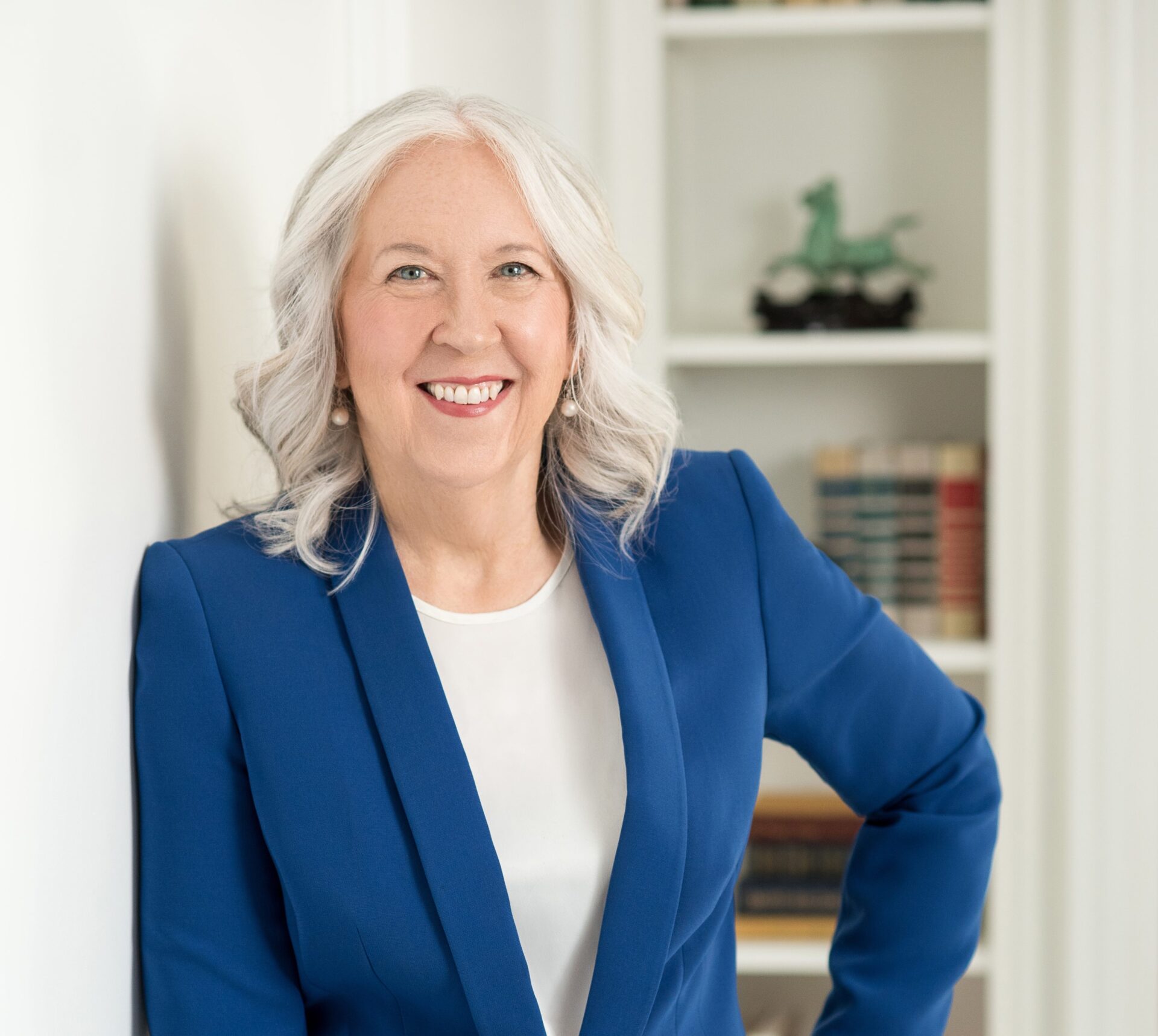 A woman with gray hair wearing a blue blazer and white shirt stands indoors, smiling, with bookshelves in the background.