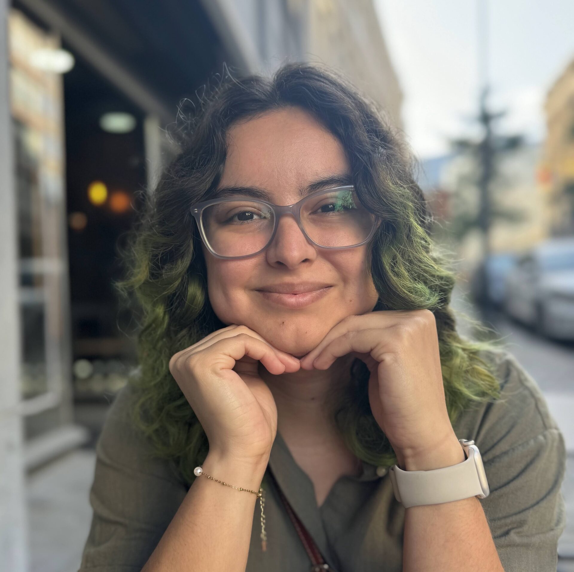A person with glasses and wavy, green-tinted hair smiles at an outdoor café, resting their chin on their hands.