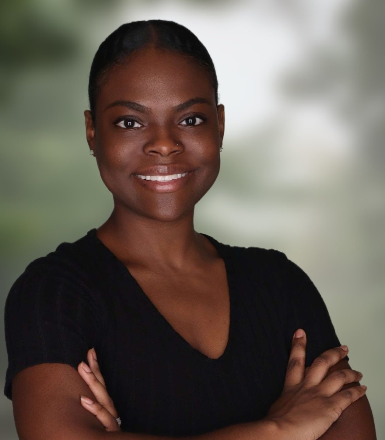 A woman with dark hair pulled back, wearing a black shirt, stands with arms crossed and smiles at the camera against a blurred background.