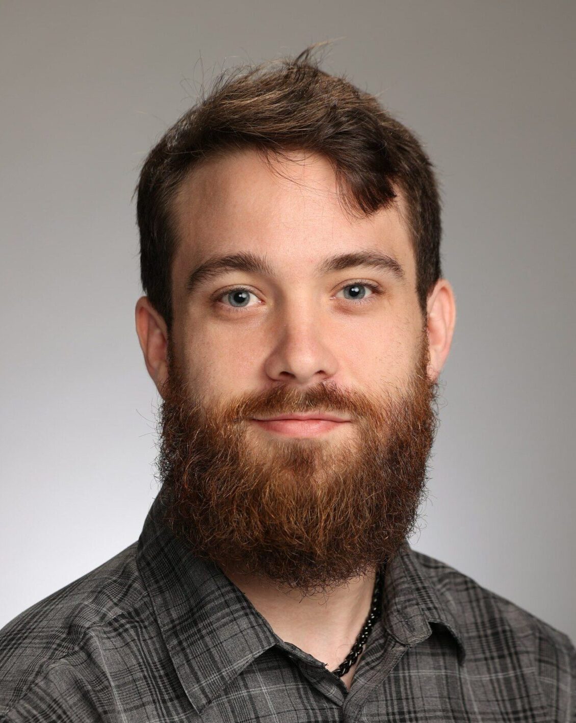 A man with a brown beard and mustache, wearing a gray plaid shirt, poses for a studio portrait against a neutral background.
