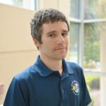 A person with short hair wearing a blue polo shirt stands indoors near large windows.