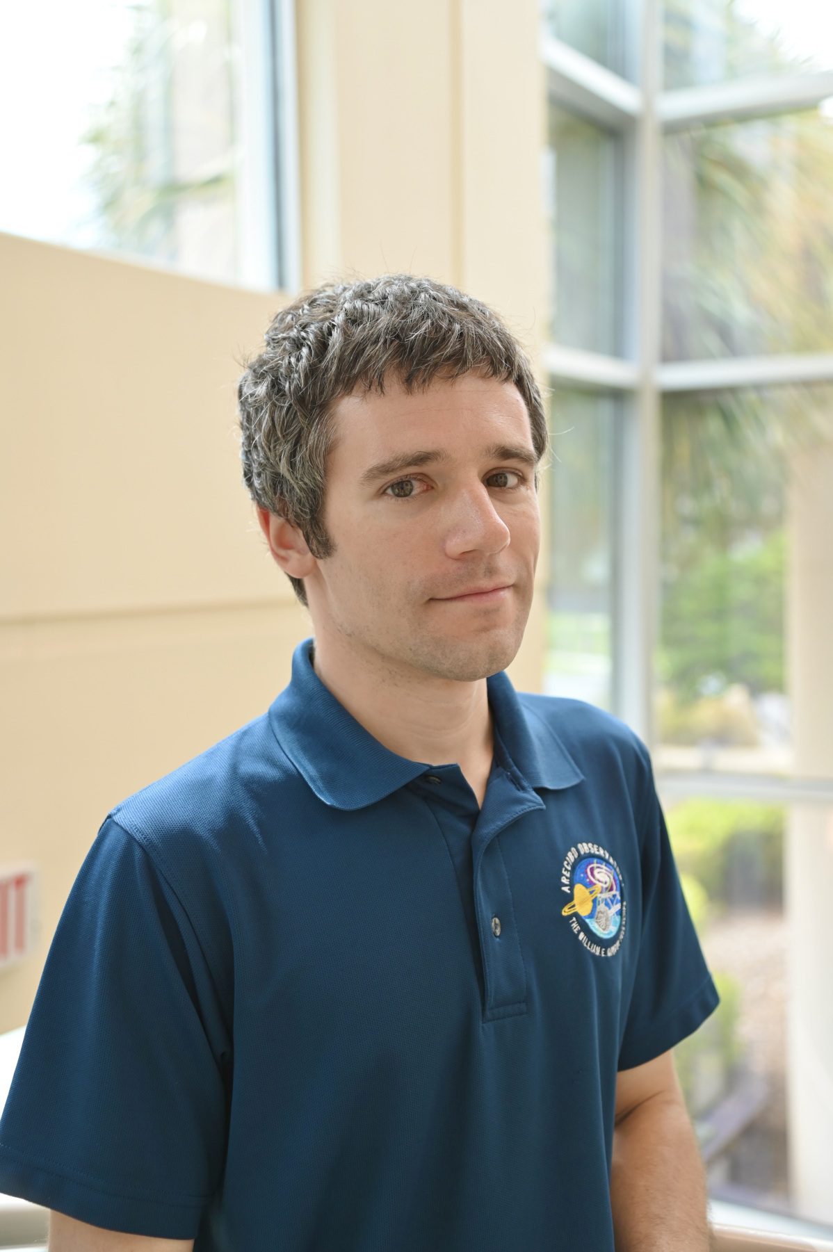 A person with short hair wearing a blue polo shirt stands indoors near large windows.