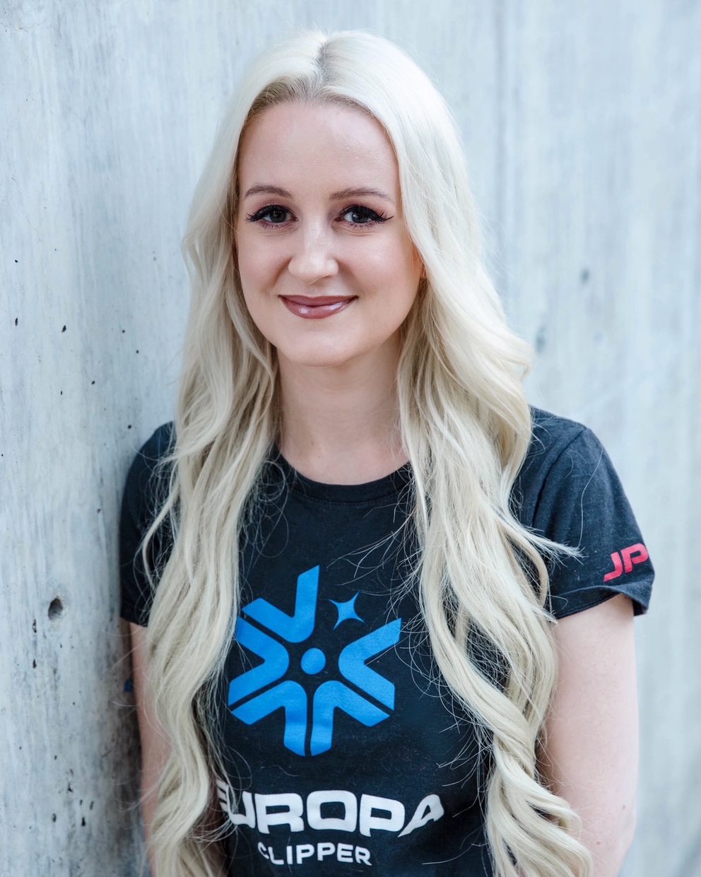 Person with long blonde hair wearing a Europa Clipper t-shirt, standing against a concrete wall.