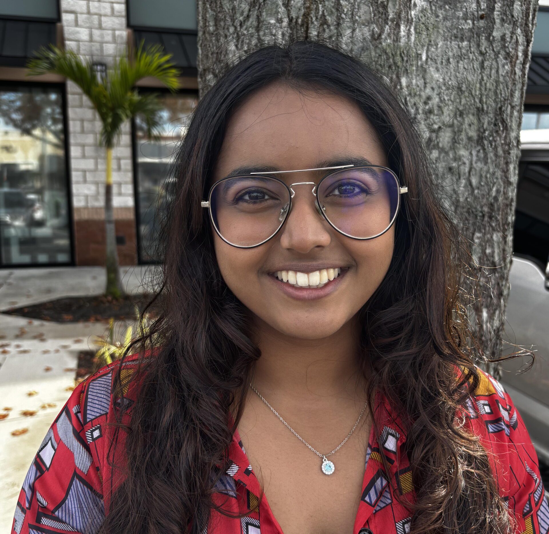 Person with long hair and glasses, wearing a red patterned shirt and a silver necklace, smiles in front of a tree and building.