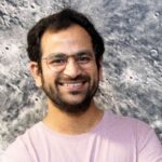 A person with glasses and a beard, wearing a pink shirt, smiles in front of a gray textured background.