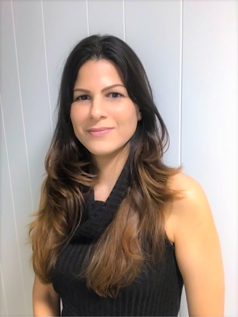 A woman with long brown hair is smiling, wearing a sleeveless black top, and standing against a light-colored wall.