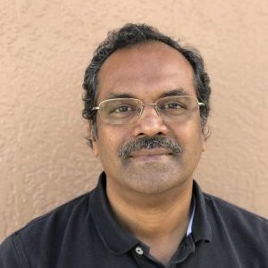 A person with glasses and a mustache, wearing a dark polo shirt, stands against a textured beige wall.