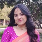 A woman with long black hair wearing a pink and orange sari, gold jewelry, and standing outdoors with greenery and trees in the background.