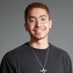 A young man with short hair and glasses smiles, wearing a black sweatshirt and a necklace with a cross pendant, against a plain gray background.
