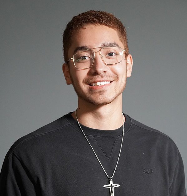 A young man with short hair and glasses smiles, wearing a black sweatshirt and a necklace with a cross pendant, against a plain gray background.