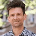 A man with short brown hair wears a patterned shirt, smiling slightly, standing outdoors with green foliage and blurred buildings in the background.