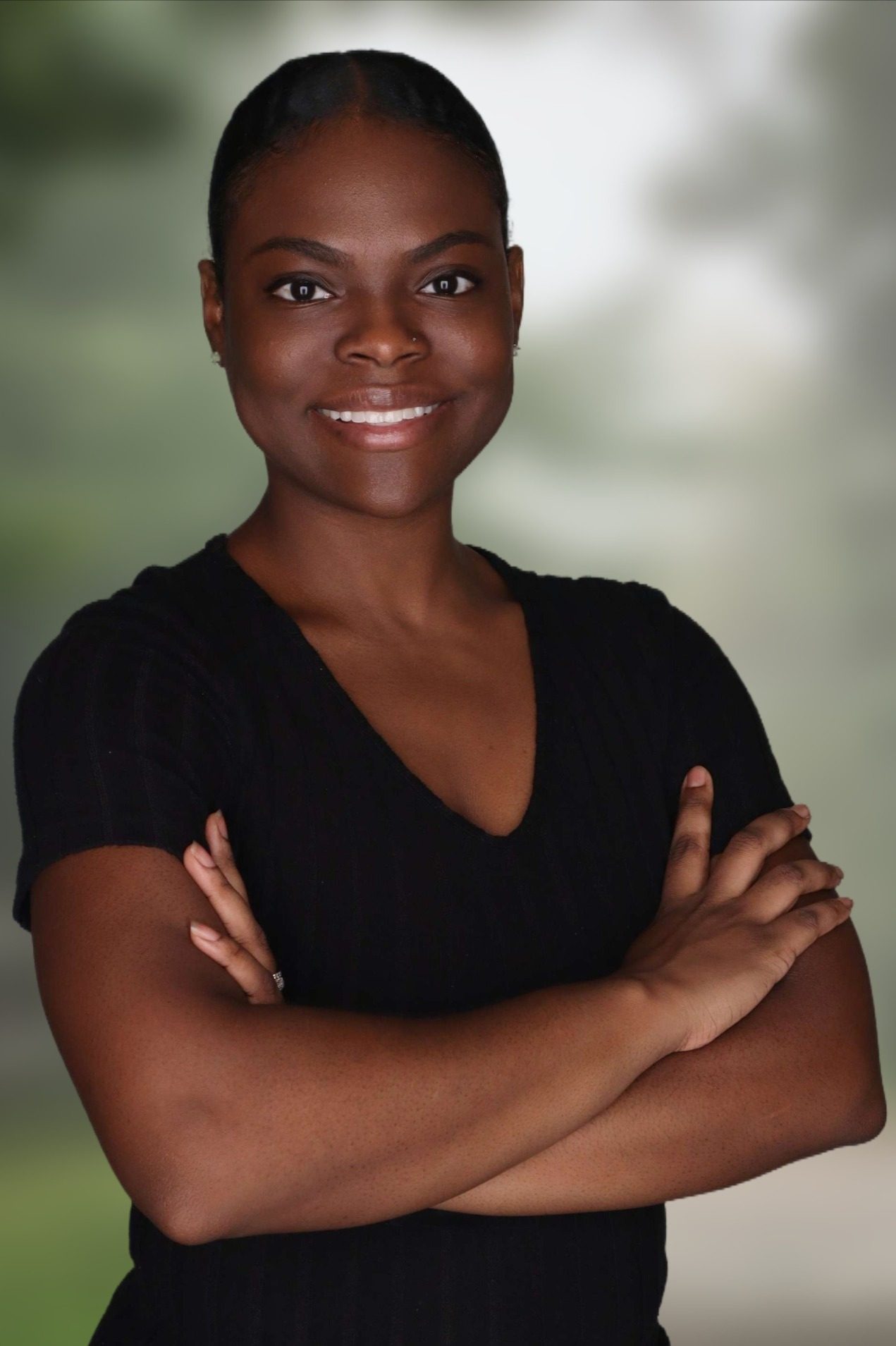 Woman standing with arms crossed, wearing a black short-sleeve top, smiling at the camera. The background is blurred with green and gray tones.