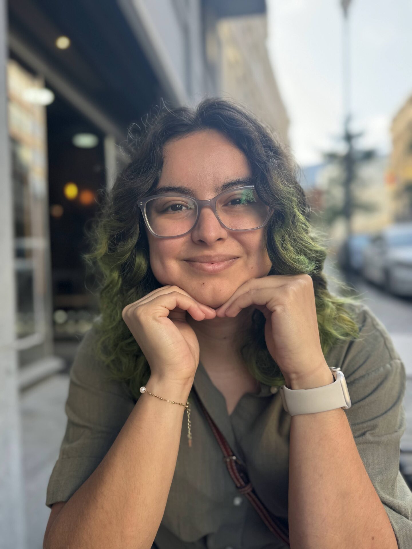 A person with glasses and wavy, green-tinted hair smiles at the camera, resting their chin on their hands while sitting outdoors at a table.