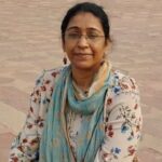 A woman with glasses and a light blue scarf sits outdoors on a stone pavement, wearing a floral-patterned top and looking at the camera.