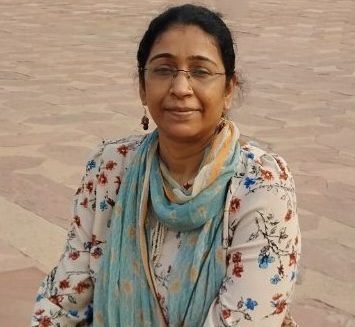 A woman with glasses and a light blue scarf sits outdoors on a stone pavement, wearing a floral-patterned top and looking at the camera.