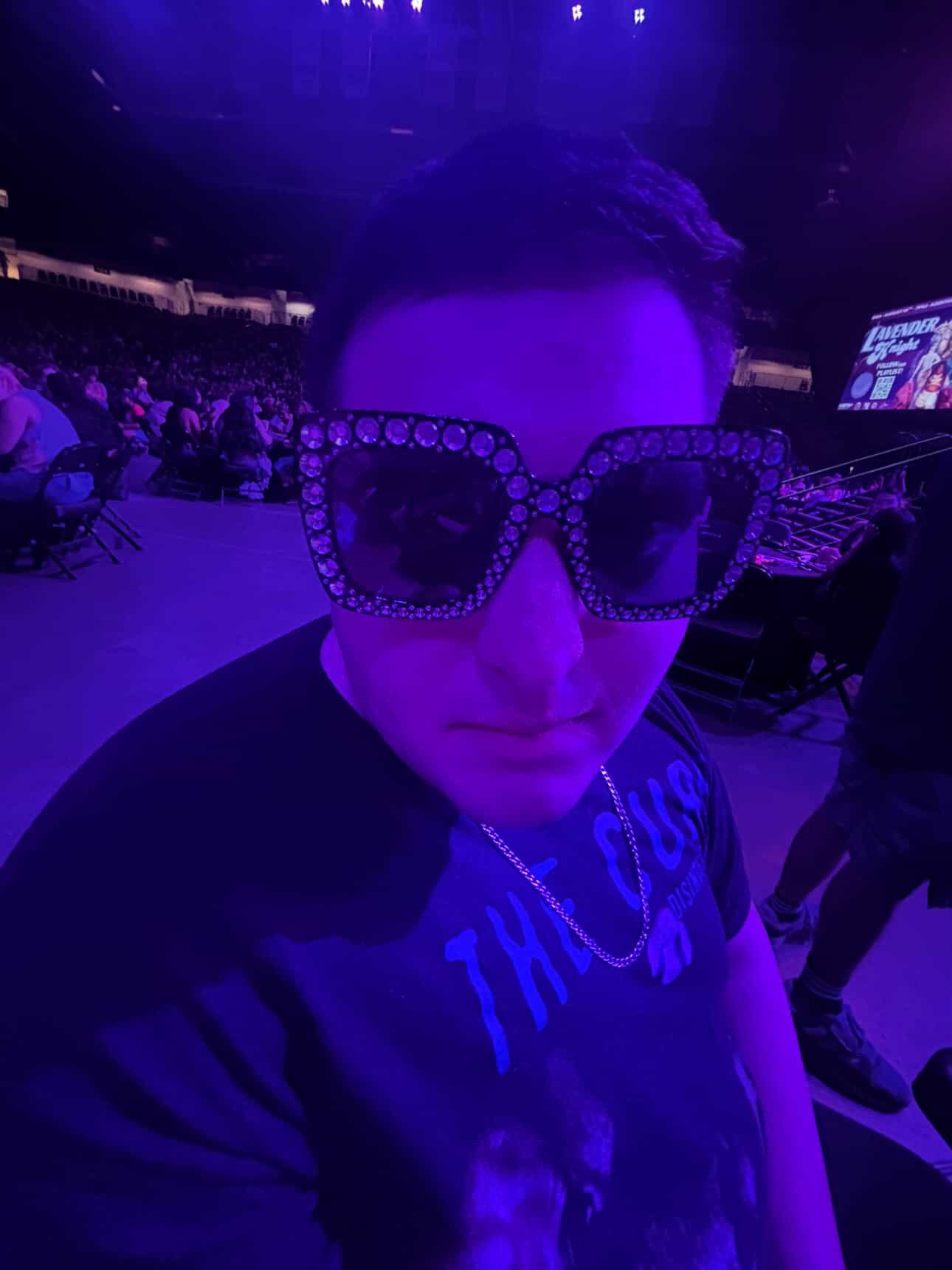 Person wearing oversized, rhinestone-studded sunglasses in an indoor arena with purple lighting. Audience and stage visible in the background.