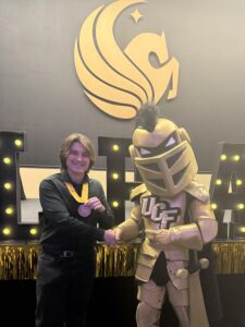 A person wearing a medal shakes hands with a knight mascot in front of a large UCF logo and decorated backdrop.
