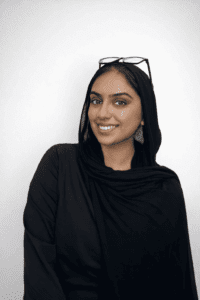 A woman wearing a black hijab and glasses resting on her head stands against a plain light background, smiling at the camera.