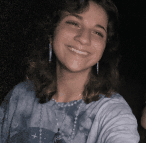 A person with shoulder-length brown hair, wearing a blue tie-dye shirt, long earrings, and a beaded necklace, smiles at the camera against a dark background.