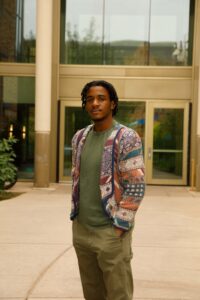 A man with short dreadlocks, wearing a patterned jacket, green t-shirt, and khaki pants, stands outside a modern building with large glass doors.