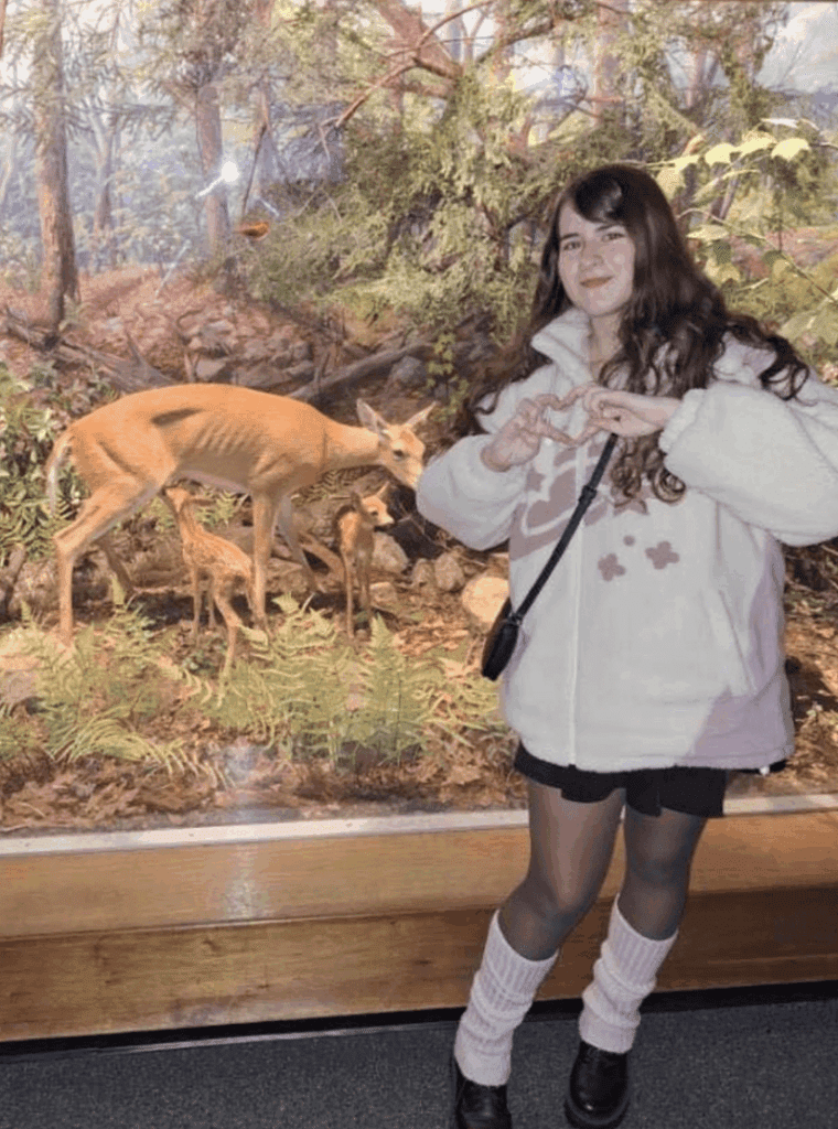 A young woman stands indoors in front of a diorama featuring a deer and fawn in a forest setting. She is wearing a light-colored jacket and making a hand gesture.
